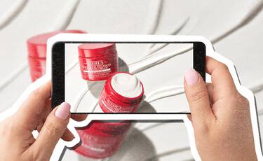 Smartphone photographing red Kiehl’s Super Multi- Corrective Cream jars against a white textured background.