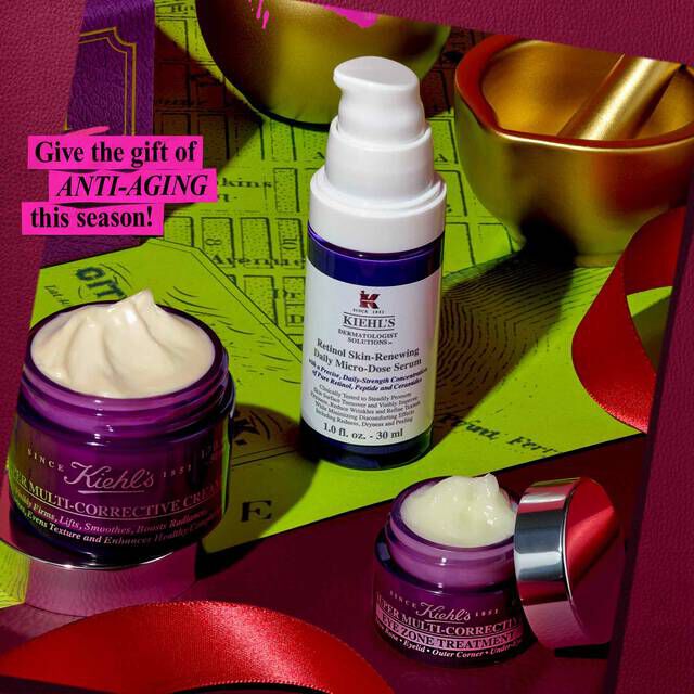 A festive graphic features three Kiehl's anti-aging products: a purple jar of "Kiehl's Super Multi-Corrective Cream," a white and purple bottle of "Kiehl's Retinol Skin-Renewing Daily Micro-Dose Serum," and a smaller purple jar of "Kiehl's Super Multi-Corrective Eye Zone Treatment." They are arranged on a green surface, with red ribbons and a gold mortar and pestle in the background. A pink text box in the upper left corner reads: "Give the gift of ANTI-AGING this season!"