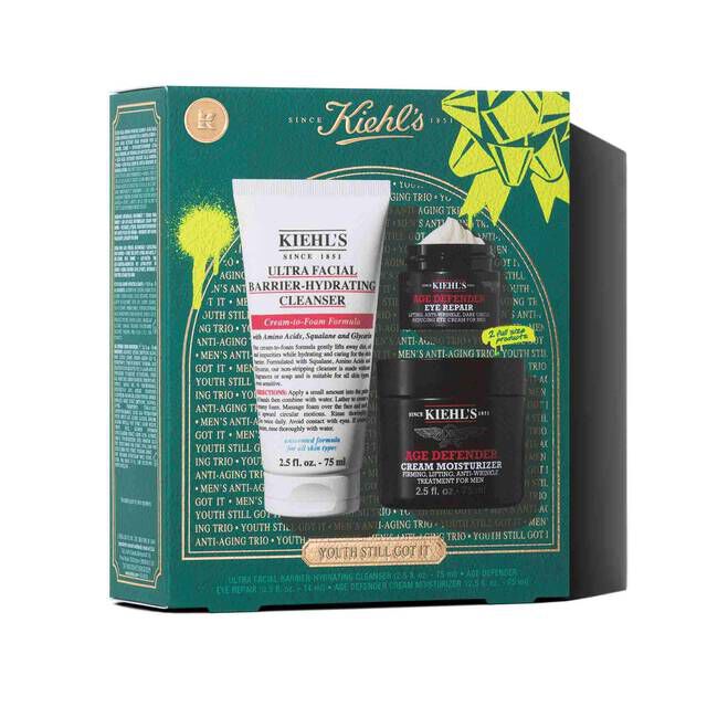 A graphic featuring a dark teal Kiehl’s gift box with white and gold accents. The box contains three products: a white tube of "KIEHL’S ULTRA FACIAL BARRIER-HYDRATING CLEANSER," a small black and red jar of "KIEHL’S AGE DEFENDER EYE REPAIR," and a larger black and red jar of "KIEHL’S AGE DEFENDER CREAM MOISTURIZER." The box is decorated with bright green graphic paint splatters and a bow design and features the text "YOUTH STILL GOT IT" at the bottom.