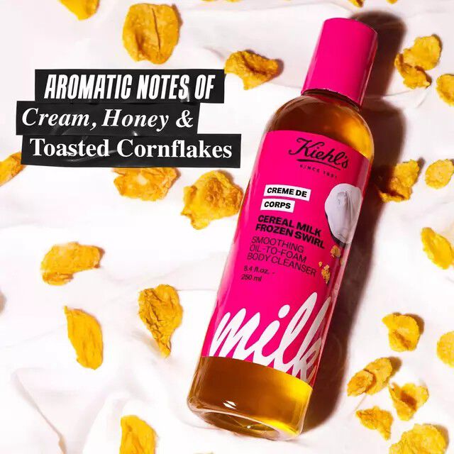 A bottle of Kiehl's Creme de Corps Cereal Milk Frozen Swirl smoothing oil-to-foam body cleanser surrounded by cornflakes, with text noting aromatic notes of cream, honey, and toasted cornflakes.​
