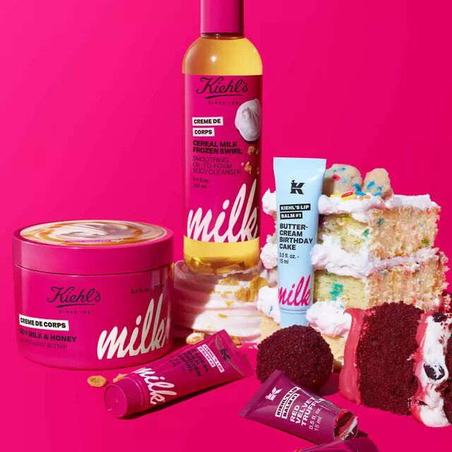 Kiehl's skincare products on a bright pink background, featuring Creme de Corps Cereal Milk body cleanser, body butter, and Lip Balm #1 tubes alongside birthday cake slices.​